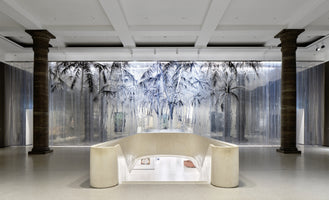 Ein heller, moderner Ausstellungsraum mit zwei dunklen, klassischen Säulen. Im Vordergrund führt eine geschwungene, cremefarbene Brüstung zu einer Treppe nach unten. Im Hintergrund eine große, transparente Wandinstallation mit einer schwarz-weißen Zeichnung eines Palmenwaldes.