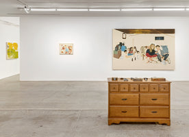 Ausstellungsansicht einer Galerie mit einer hölzernen Kommode im Vordergrund. An der weißen Wand dahinter hängen zwei Gemälde: ein großes, das eine Gruppe von Menschen zeigt, und ein kleineres, abstraktes. Links im Hintergrund ist ein weiteres Gemälde zu sehen.