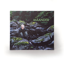 Das quadratische Buch MÄANDER von moki. Das Cover ist ein Gemälde, das eine Person mit blonden Haaren in einem dunklen Fluss zwischen moosbewachsenen Felsen zeigt. Das Buch liegt auf einer weißen Fläche.