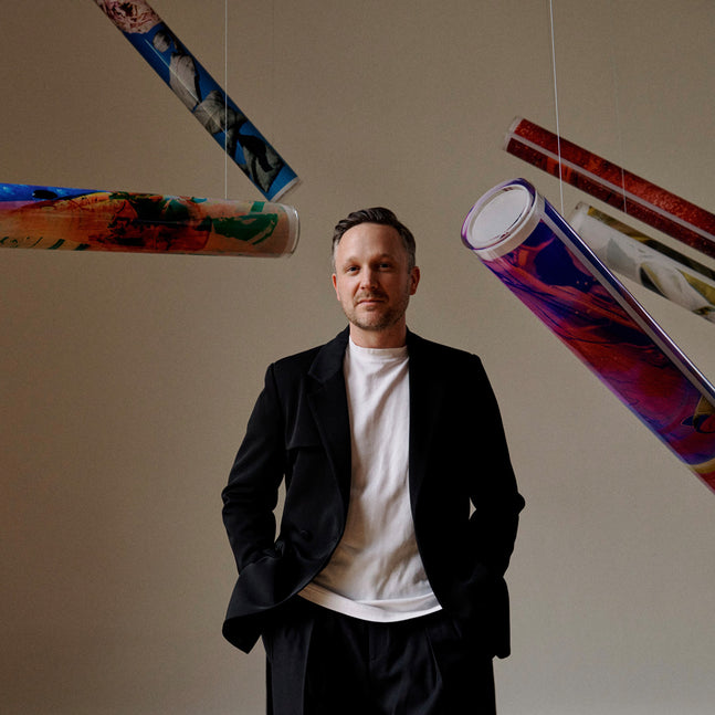 Ein Mann in einem schwarzen Sakko und einem weißen T-Shirt steht mit den Händen in den Hosentaschen vor einer hellen Wand. Er blickt direkt in die Kamera. Um ihn herum hängen mehrere bunte, zylindrische Objekte an dünnen Fäden von der Decke, die wie aufgerollte Kunstwerke aussehen.
