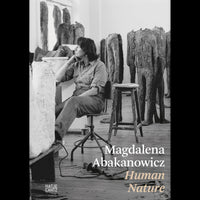 Eine Schwarz-Weiß-Fotografie der Künstlerin Magdalena Abakanowicz, die nachdenklich in ihrem Atelier sitzt, umgeben von ihren großen, kopflosen Skulpturen.