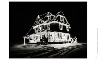 Eine Schwarz-Weiß-Fotografie eines großen, mehrstöckigen Hauses bei Nacht, das vollständig mit leuchtenden Weihnachtslichtern geschmückt ist. Das Haus steht in einer schneebedeckten Landschaft, und im Vordergrund sind Reifenspuren im Schnee sichtbar.