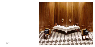 Eine Fotografie eines surrealen Esszimmers mit holzgetäfelten Wänden und einem schwarz-weiß karierten Boden. Ein langer Tisch ist mit einem weißen Tischtuch für zwei Personen gedeckt. Anstelle der Gäste stehen zwei alte Röhrenfernseher auf Stühlen und blicken auf die leeren Plätze am Tisch.