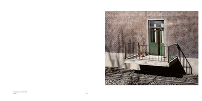 Ein kleiner Betonbalkon mit schwarzem Metallgeländer ragt aus einer Backsteinwand direkt über Eisenbahngleisen. Eine grüne, verglaste Tür steht offen zum Balkon, auf dem eine Topfpflanze steht. Helles Sonnenlicht wirft harte, dunkle Schatten.