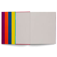 Ein von oben fotografiertes, aufgeschlagenes Buch mit rotem Buchrücken. Links ist der Einband mit vertikalen Streifen in Lila, Orange, Blau und Gelb zu sehen, rechts die beiden leeren, hellgrauen Innenseiten.