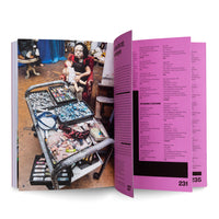 Ein aufgeschlagenes Kunstbuch, das auf der linken Seite ein Foto eines Ateliers mit einer großen Puppe im Sessel und einem Wagen voller Künstlerbedarf zeigt und auf der rechten, pinkfarbenen Seite eine Werkliste in schwarzer Schrift.