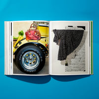 Ein aufgeschlagenes Buch liegt auf einer leuchtend blauen Oberfläche, von oben fotografiert. Die linke Buchseite zeigt das Foto eines gelben Autos mit einer roten Handtasche auf dem Kotflügel, die rechte Seite zeigt ein schwarz-weiß gepunktetes Kleid vor einem Hintergrund aus Notenblättern.