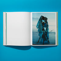 Ein aufgeschlagenes Buch auf einem leuchtend blauen Untergrund. Die linke Seite ist leer, während die rechte Seite eine Fotografie einer alten Großformatkamera auf einem Stativ zeigt, die am Ufer eines Gewässers steht. Ein blaues, durchsichtiges Tuch weht um die Kamera, und im Hintergrund sind neblige Berge und ein fliegender Vogel zu erkennen.