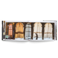 Ein aufgeschlagenes Buch zeigt eine Fotografie, die sich über eine Doppelseite erstreckt. Sie zeigt eine sonnendurchflutete Arkade mit hohen Steinbögen, in denen lange, durchscheinende Vorhänge in Gold- und Weißtönen hängen. Ein Mann in einem schwarzen T-Shirt geht an der Arkade vorbei.